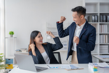 Two young Asian business people successful excited raised hands rejoicing with a laptop computer in a modern office.