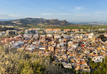 Fototapeta na wymiar Buildings, houses and streets in city, aerial view. View of rooftops and streets of city of Sagunto in Spain. Town against backdrop of mountains. Roofs of houses and roofs from side of Sagunto Castle.