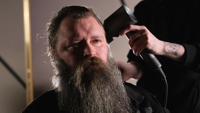 a young stylist does styling with a hair dryer to a bearded man sitting in a barber chair.