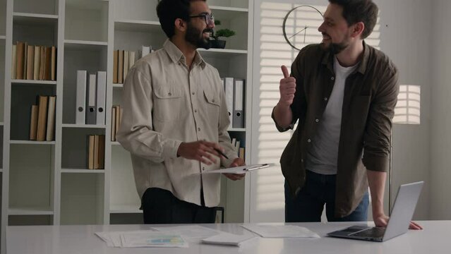 Two multiracial businessmen partners smiling men discuss business project on laptop show thumb up handshaking partnership deal agreement financial benefit at office meeting handshake Generative AI