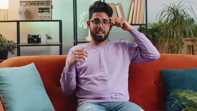 I dont know what to say. Confused indian handsome man feeling embarrassed about ambiguous question, having doubts, no idea, being clueless and uncertain at home. Hindu guy sits on sofa in living room