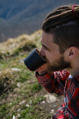 Guy in red plaid shirt with dreadlocks and beard sitting on vacation during mountain hike and drinking hot tea from mug. Portrait of male tourist, side view. Concept of travel and active lifestyle.