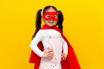 little asian girl in superman costume and mask, korean child in superhero cape smiling on yellow isolated background