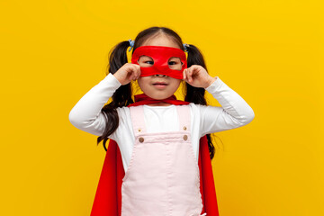little asian girl in superman costume and mask, korean child in superhero cape smiling on yellow isolated background