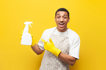 guy african american cleaner in apron holds gloves and detergent on yellow isolated background, cleaning service
