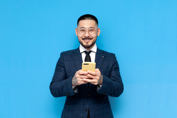 successful asian businessman in suit use smartphone and smile on blue isolated background, korean entrepreneur