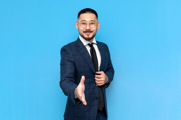 successful asian businessman in suit and glasses gives handshake on blue isolated background