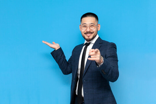 hey you, successful asian businessman in suit and glasses points at you on blue isolated background