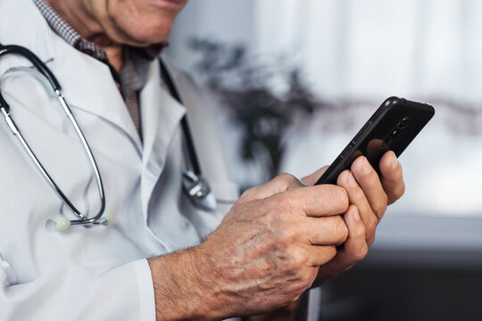 Mature doctor wearing uniform typing in smart phone holding it in hands closeup side view.