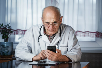 Senior doctor holding smartphone and texting message video calling for telemedicine consultation online. Therapeutist with phone in hands in medical office.