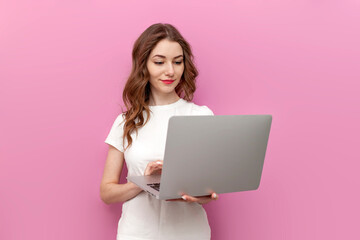 young cute woman in white t-shirt uses laptop on pink isolated background, woman freelancer types...