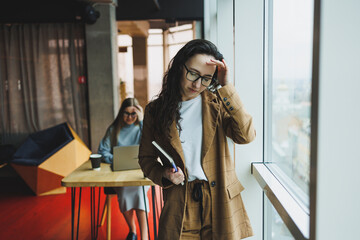 Fototapeta premium A successful woman in glasses works in the office on the background of colleagues. Modern working day in the office. Businesswoman makes notes in a notebook