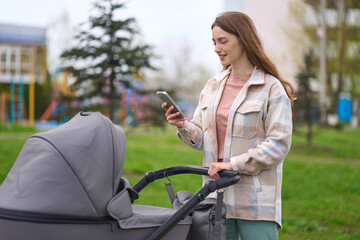 Fototapeta na wymiar Happy young mother checking smartphone while walking with her newborn child in baby stroller in park