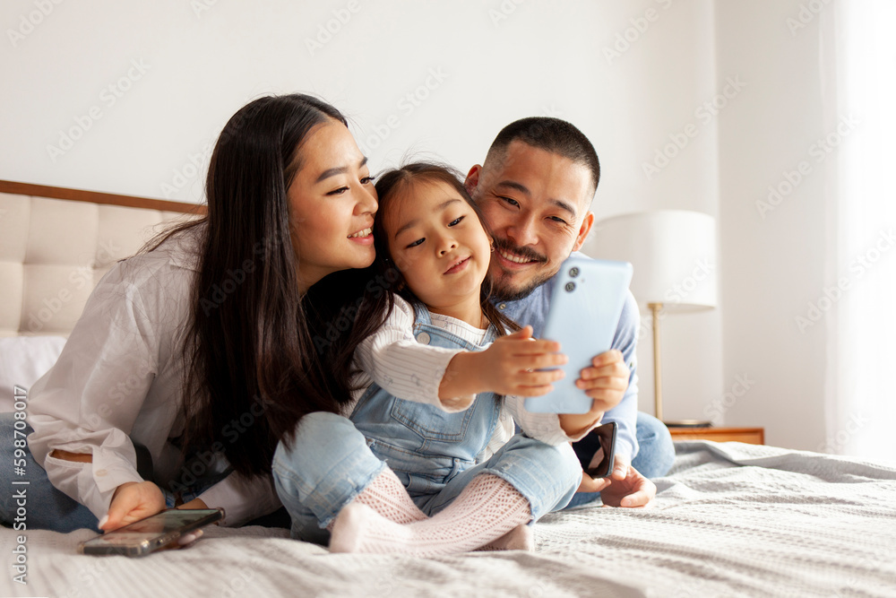 Wall mural asian family sit on the bed and communicate via video call, little korean girl with mom and dad take