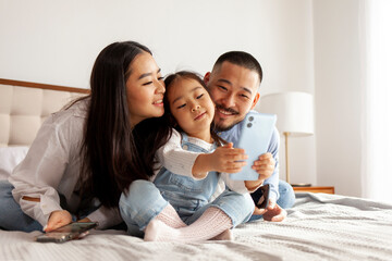asian family sit on the bed and communicate via video call, little korean girl with mom and dad...