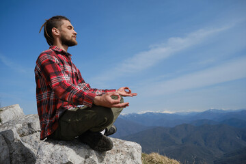 Naklejka na ściany i meble Guy traveler sitting on big rock in mountains and enjoying views of nature of Caucasus. Young man with dreadlocks meditates and does yoga outside. Side view. Concept of travel and active lifestyle.
