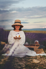 A woman in a white dress sits in the middle of a lavender field and holds fragrant tea in her hands. Blooming lavender. Picnic in Provence style.