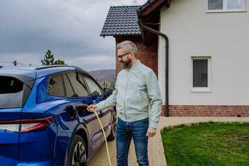Mature man holding power supply cable and charging his electric car.