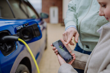 Close up of man and his daughter holding smartphone, checking charging of his electric car.