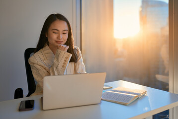 Young happy Asian professional business woman using laptop technology in company office, looking at computer watching webinar, feeling satisfied with corporate results management sitting Generative AI - obrazy, fototapety, plakaty