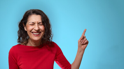 Happy and smiling mature adult brunette caucasian female with red sweater points away with finger over pastel blue background with copy space