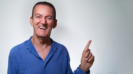Funny and smiling mischievous mature adult caucasian male with blue shirt points away with finger over white grey background with copy space