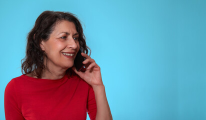 Happy and smiling mature adult brunette caucasian female with red sweater speaks on smartphone over pastel blue background with copy space