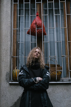Young male model brutal man wearing long hair and black clothes standing near window with bars and hanging vegetables on city street