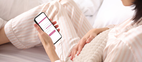 Young woman holding mobile phone with open menstrual calendar on screen in bedroom