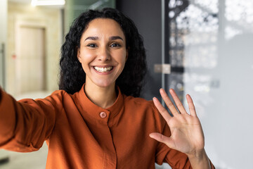 Beautiful business woman with curly hair wearing looking at smartphone camera, waving her hand in...