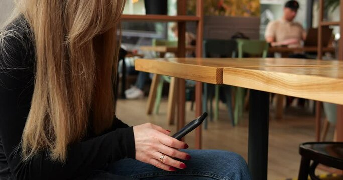Unrecognizable young woman in coffee shop waiting for order with phone on knees. Faceless female with long brown hair sits at empty table type on smartphone, cars and people in street in Generative AI