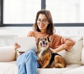  Happy young woman spending time with her pet dog at home sitting on couch and browsing in  tablet.