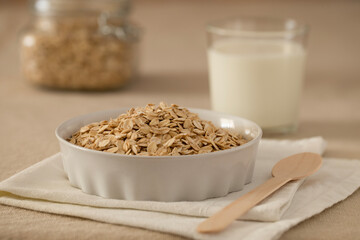 Oat flakes, cereal in a white ceramic bowl on a linen tablecloth on the table, a glass of milk, a spoon and a glass jar of oatmeal