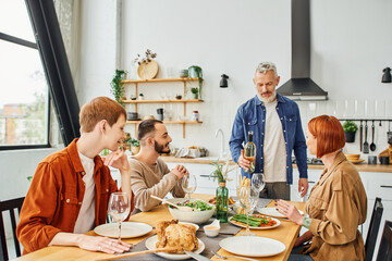 smiling man standing with wine bottle near family and tasty supper served in kitchen. 