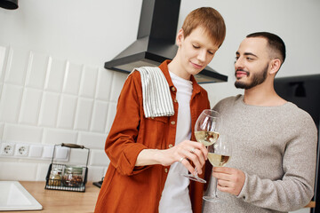 bearded gay man clinking wine glasses with smiling redhead boyfriend kitchen. 