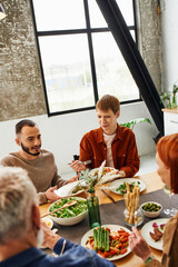 bearded gay man talking near redhead boyfriend and blurred parents during family supper in kitchen. 