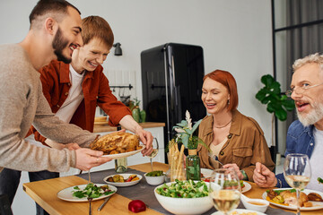 joyful gay couple serving grilled chicken near happy parents in kitchen. 