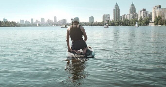 Man sailing away from pier on electric surfboard, close up. Active lifestyle on morning modern cityscape background. Lift Foil Electric Hydrofoil Surfboard. Using electrofoil in city blue river water