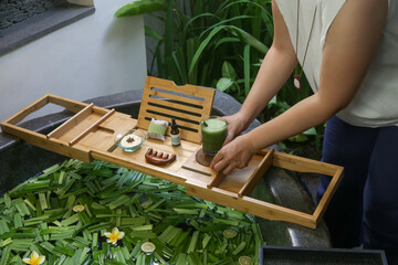 Tub full of water and herbs with wooden bathtub tray with various cosmetic products, relaxation and wellbeing concept of lifestyle	