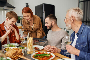 redhead woman holding family photo album near son with gay partner during delicious supper in kitchen. 