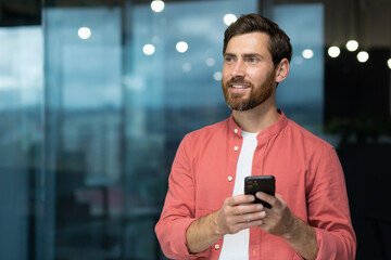 Portrait of a young freelancer man manager standing in the office and using a mobile phone. He looks to the side with a smile, waiting for a meeting.