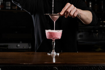 Bartender is pouring pink Clover club alcoholic cocktail in the glass. Bartender mixes egg white,...