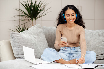 Positive young Arab woman sits on the sofa and smiles, writes in a notebook and listens to music on headphones. Woman holds a phone in her hands checks data with a laptop listen to her favorite song