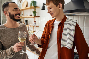 redhead man clinking wine glasses with happy bearded boyfriend in kitchen. 