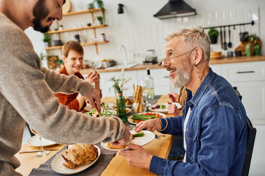 smiling bearded man looking at gay man serving chicken during supper with family in kitchen. 
