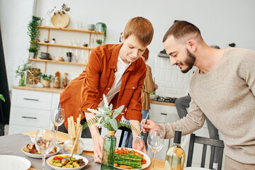 gay man holding fork near boyfriend holding grilled asparagus near table with family supper in...
