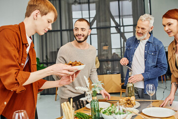 redhead gay man holding grilled vegetables near boyfriend and happy parents in kitchen. 