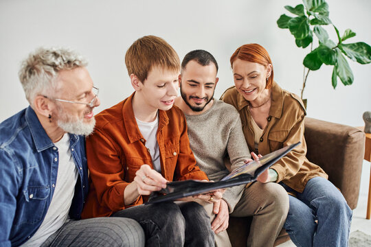 redhead gay man looking at photo album together with boyfriend and joyful parents. 