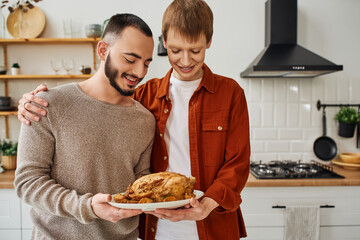 happy gay man embracing boyfriend while holding grilled chicken in kitchen. 
