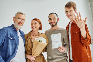 happy gay man showing wedding ring near parents with flowers and boyfriend with wedding invitation. 